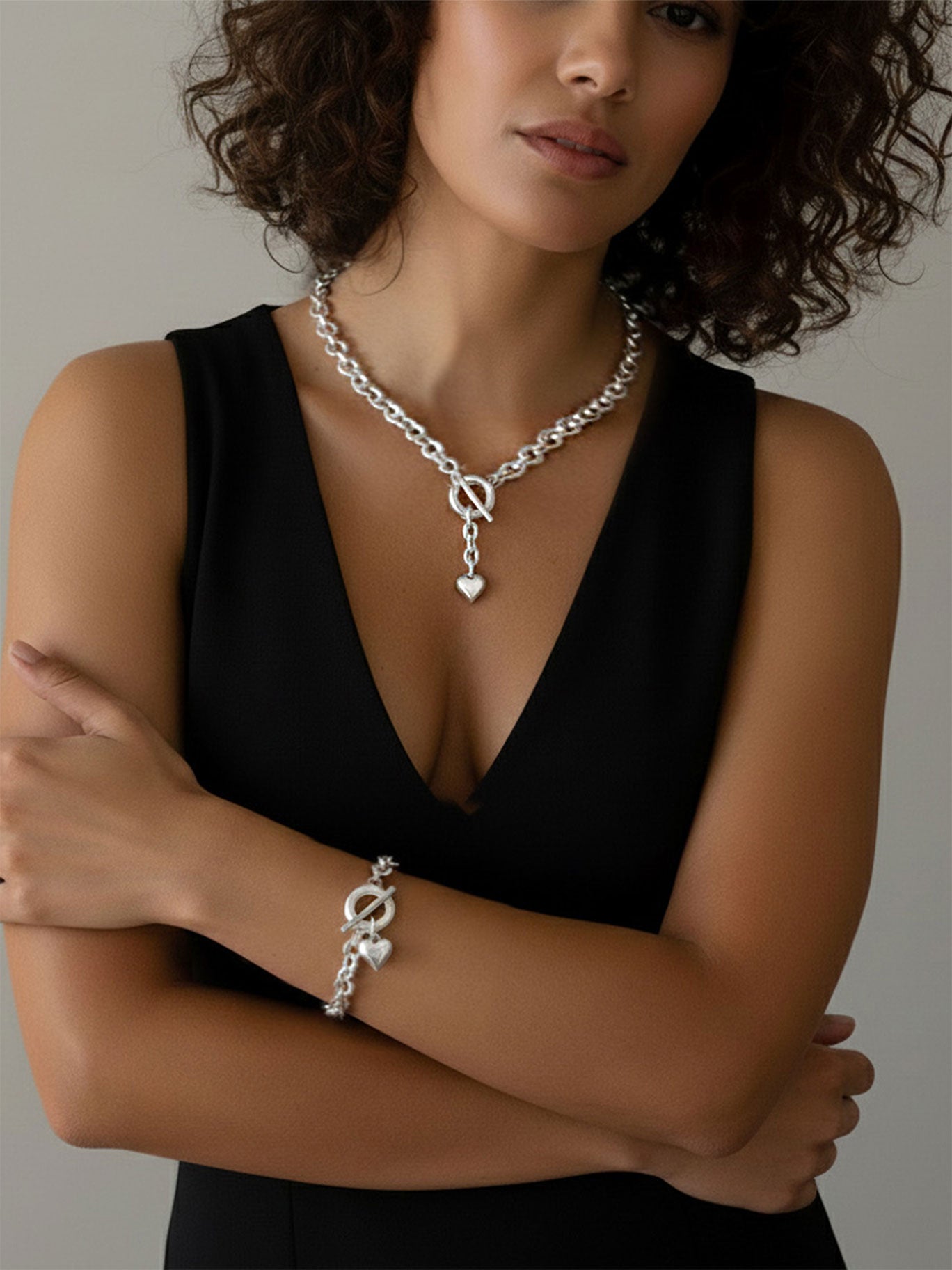 Frau mit lockigem Haar trägt Schmuck-Set K306+A306 chuznky Ketten-Collier mit Herzanhänger und passendem Armband mit Knebelverschluss, ideal als Geschenk oder elegantes Accessoire.